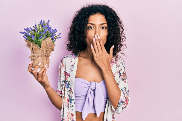 Nuori latino tyttö tilalla laventeli kasvi peittää suun käsin, järkyttynyt ja pelkää virhe. yllättynyt ilme  - Valokuva, kuva