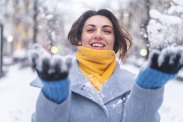 Eine junge Frau in Mantel und gelbem Schal auf dem Stadtboulevard lächelt am Abend fröhlich, wirft Schnee in die Kamera - Foto, Bild