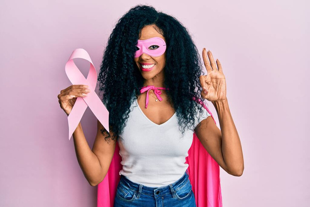 中世アフリカ系アメリカ人女性を身に着けていますスーパーヒーロー衣装を保持ピンクの癌リボンを持っています指でokサインを行います,笑顔優しいジェスチャー優れたシンボル  - 写真・画像