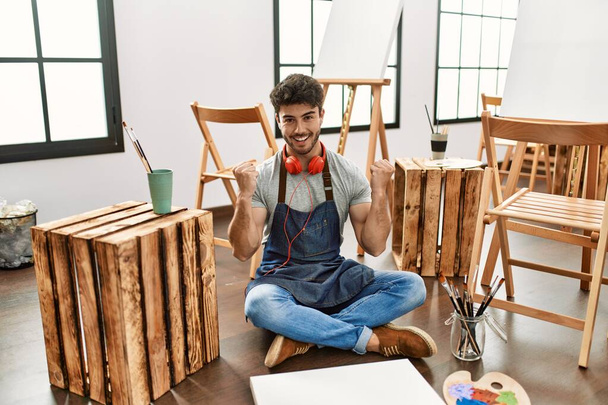 Sanat stüdyosunda oturan İspanyol genç adam şaşkınlık ve şaşkınlık içinde kollarını kaldırıp gözleri açık bir şekilde başarıyı kutluyor. Kazanan konsept.  - Fotoğraf, Görsel
