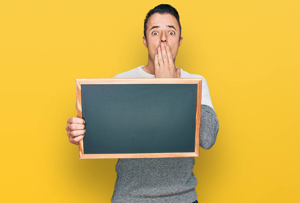 Handsome young man holding blackboard covering mouth with hand, shocked and afraid for mistake. surprised expression  - Fotoğraf, Görsel