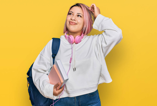 Spaanse vrouw met roze haar dragen student rugzak en koptelefoon glimlachen vol vertrouwen aanraken haar met de hand omhoog gebaar, poseren aantrekkelijk en modieus  - Foto, afbeelding