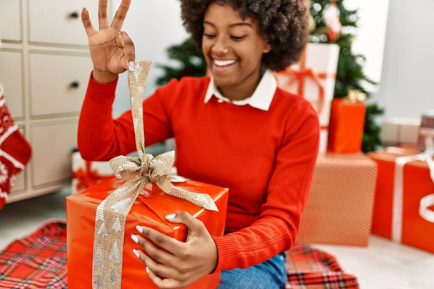 Молода афроамериканська жінка, яка розвантажує подарунок, сидячи біля христми вдома. - Фото, зображення