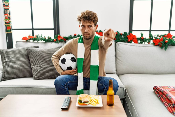Młody Hiszpan futbol chuligan trzymając piłkę w domu wskazując palcem na aparat i do ciebie, pewny siebie gest wygląda poważnie  - Zdjęcie, obraz