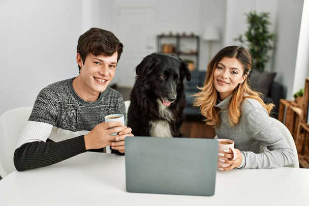 若いですcaucasianカップル使用してノートパソコンとコーヒーを飲みますテーブルの上に座って犬と自宅で. - 写真・画像