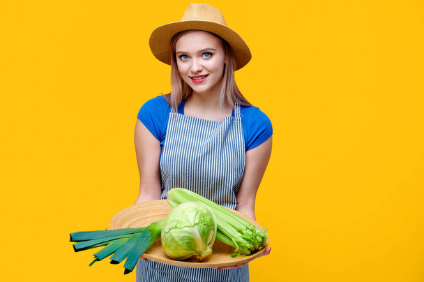 Женщина-садовница в соломенном фартуке в шляпе держит овощи сельдерей капусты и лук-порей на деревянном подносе на желтом фоне - Фото, изображение