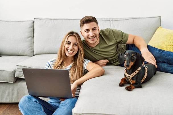 Jeune couple hispanique souriant heureux en utilisant un ordinateur portable assis sur le canapé avec chien à la maison. - Photo, image