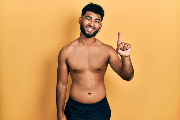 Arabische man met baard draagt zwemkleding zonder shirt te tonen en te wijzen met vinger nummer een, terwijl hij vol vertrouwen en gelukkig glimlacht.  - Foto, afbeelding
