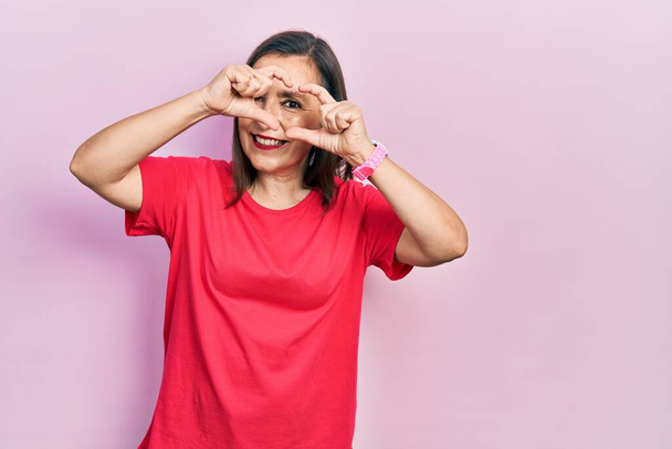 Middle age hispanic woman wearing casual clothes doing heart shape with hand and fingers smiling looking through sign  - Valokuva, kuva