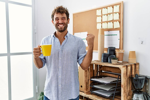 Mladý pohledný muž pije šálek kávy v kanceláři křičí hrdě, slaví vítězství a úspěch velmi vzrušený se zvednutou paží  - Fotografie, Obrázek