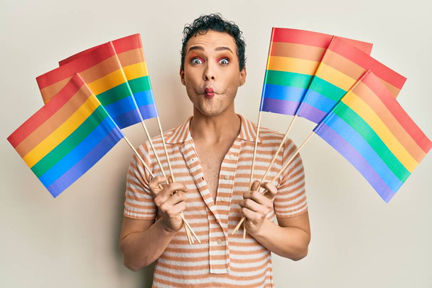 Hombre guapo usando maquillaje sosteniendo banderas de arco iris lgbtq haciendo cara de pez con boca y ojos entrecerrados, loco y cómico.  - Foto, Imagen