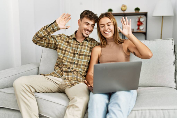 Молодая латиноамериканская пара с видеозвонком с помощью ноутбука сидит на диване дома. - Фото, изображение