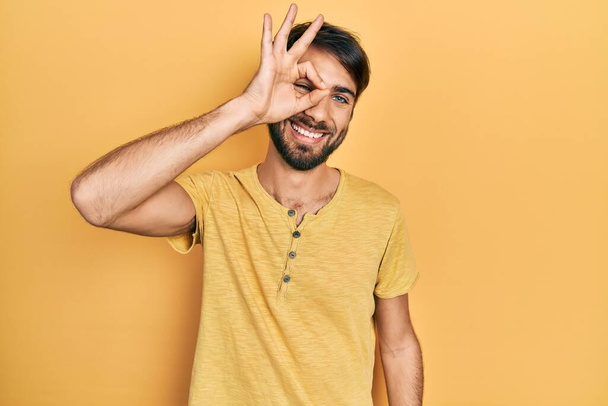 Молодий іспаномовний чоловік в повсякденному одязі посміхається щасливим роблячи знак з рукою на око дивиться крізь пальці
  - Фото, зображення