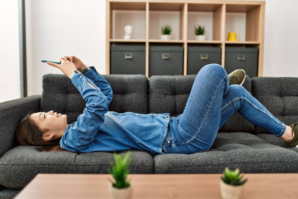 Giovane ragazza cinese utilizzando smartphone sdraiato sul divano a casa. - Foto, immagini