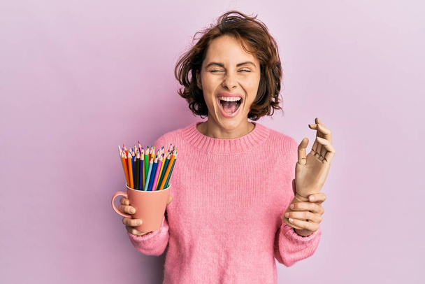 Jeune femme brune tenant une petite main de mannequin en bois et des crayons souriant et riant fort à haute voix parce que drôle blague folle.  - Photo, image
