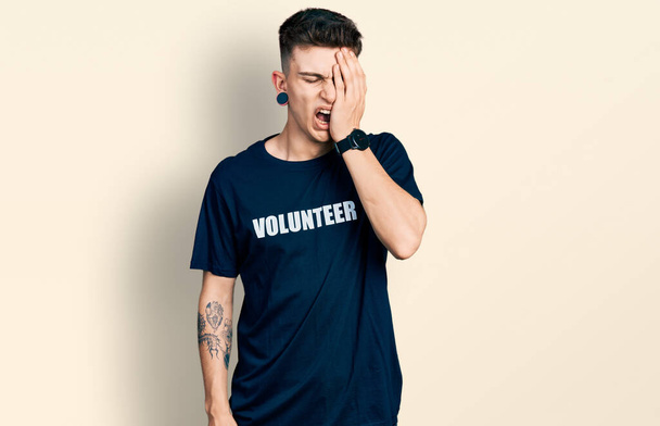 Молодий білий хлопчик з розширенням вух носить волонтерську сорочку, що позіхає втомлене обличчя, око і рот рукою. болить обличчя від болю
.  - Фото, зображення