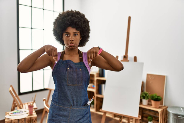 Молодая африканская американка с афроволосами в художественной студии, указывающая на грустный и расстроенный вид, указывающая направление пальцами, несчастная и подавленная.  - Фото, изображение