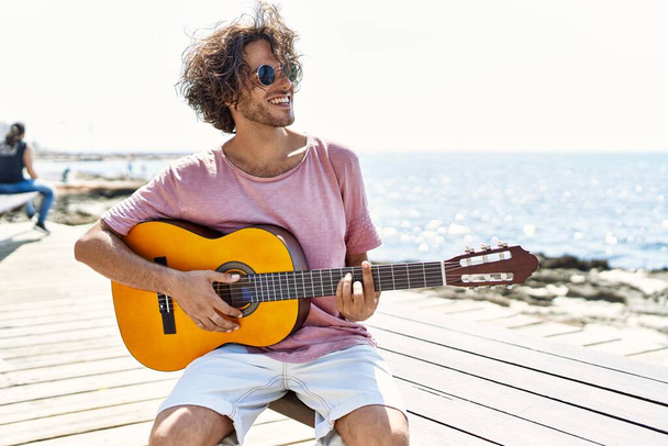 Young hispanic man playing classical guitar sitting on  bench at the beach. - Zdjęcie, obraz
