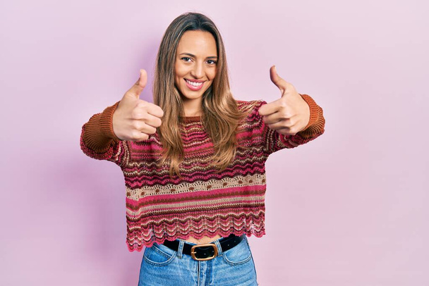 美しいヒスパニック系の女性は、手で積極的なジェスチャーを行うことを承認ヒッピーセーターを着て,親指アップ笑顔と成功のために幸せ.勝者のジェスチャー.  - 写真・画像