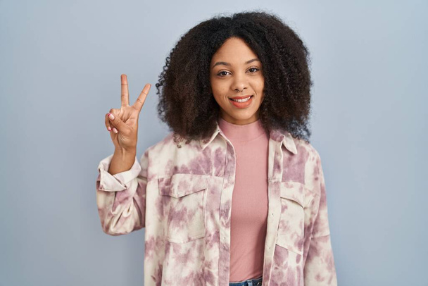 自信を持って幸せな笑顔ながら、青の背景に立って指の数2で指摘している若いアフリカ系アメリカ人女性.  - 写真・画像