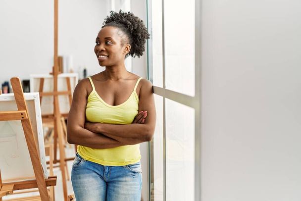 Νεαρή Αφρικάνα Αμερικανίδα καλλιτέχνιδα χαμογελαστή χαρούμενη στέκεται με σταυρωμένα τα χέρια στο στούντιο τέχνης. - Φωτογραφία, εικόνα