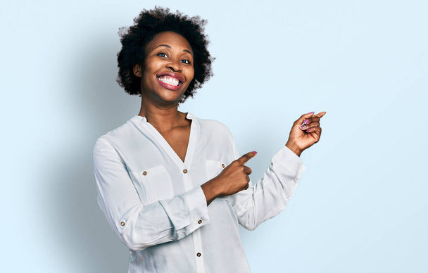 Afrikkalainen amerikkalainen nainen afro hiukset yllään rento valkoinen t paita hymyilee ja katsot kameraa osoittaa kahdella kädellä ja sormella sivulle.  - Valokuva, kuva