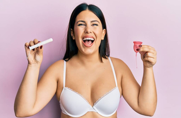 Krásná brunetka žena držící menstruační pohár ve spodním prádle překvapená nápadem nebo otázkou ukazující prstem se šťastnou tváří, číslo jedna  - Fotografie, Obrázek
