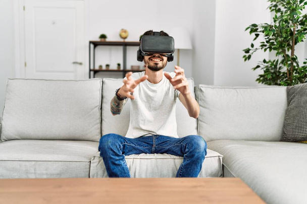 Jeune homme hispanique jouant à un jeu vidéo en utilisant des lunettes de réalité virtuelle à la maison. - Photo, image