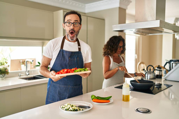 Couple d'âge moyen cuisine des aliments méditerranéens à la maison effrayé et choqué par la surprise et l'expression étonnante, la peur et le visage excité.  - Photo, image