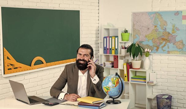brutal bearded man work in classroom and hols smartphone. prepare for online exam. college lecturer on lesson. back to school. formal education. conversation. happy mature teacher speak on phone - Fotografie, Obrázek