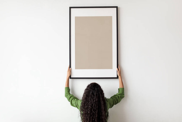 Cartel en blanco colgante femenino irreconocible en el marco en la pared interior - Foto, Imagen