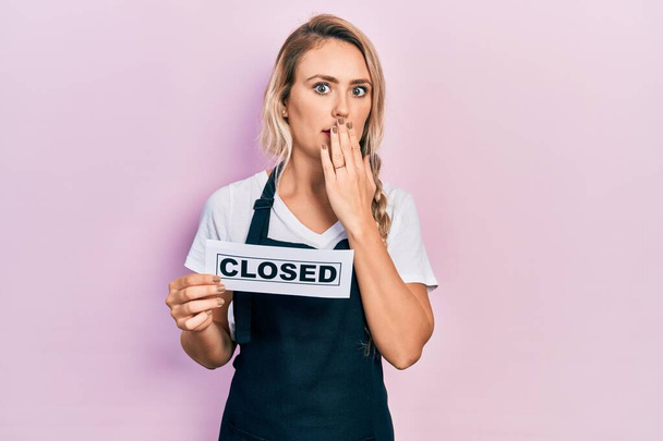Beautiful young blonde woman wearing waitress apron holding closed banner covering mouth with hand, shocked and afraid for mistake. surprised expression  - Fotografie, Obrázek