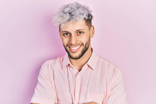 Joven hombre hispano con el pelo teñido moderno con camisa rosa casual cara feliz sonriendo con los brazos cruzados mirando a la cámara. persona positiva.  - Foto, imagen