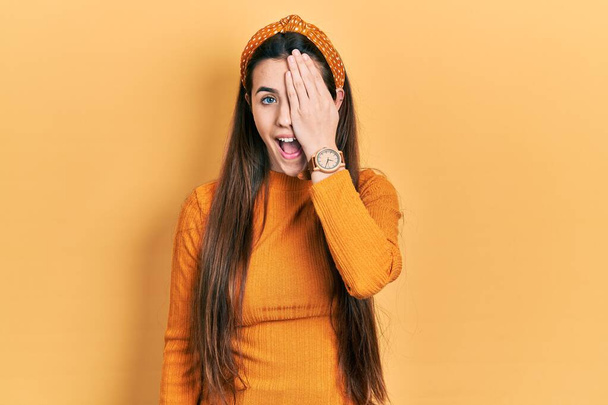 Młoda brunetka nastolatka ubrana w luźny żółty sweter zakrywający jedno oko ręką, pewny siebie uśmiech na twarzy i niespodziewane emocje.  - Zdjęcie, obraz