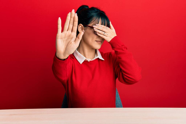 Young brunette woman with bangs wearing glasses sitting on the table covering eyes with hands and doing stop gesture with sad and fear expression. embarrassed and negative concept.  - Foto, afbeelding