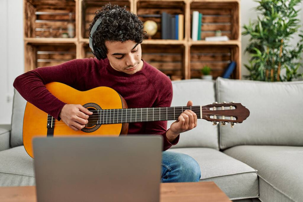 Young hispanic man having online classical guitar lesson sitting on the sofa at home. - Photo, Image