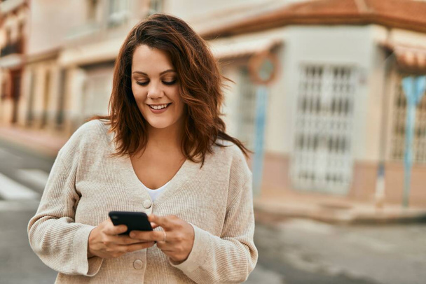 Genç İrlandalı artı cüsseli kız şehirde akıllı telefon kullanarak mutlu gülümsüyor.. - Fotoğraf, Görsel