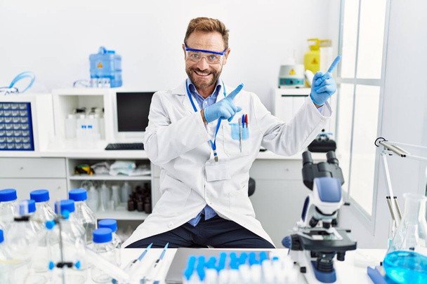 Hombre de mediana edad que trabaja en el laboratorio científico sonriendo y mirando a la cámara apuntando con dos manos y dedos hacia un lado.  - Foto, Imagen