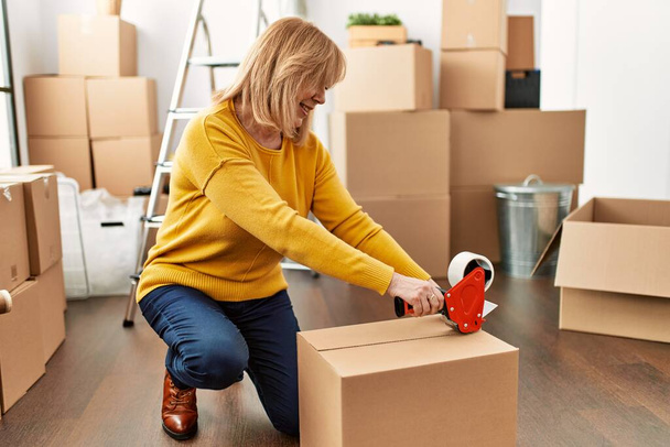 Blondynka w średnim wieku pakująca pudełko z taśmą w nowym domu. - Zdjęcie, obraz
