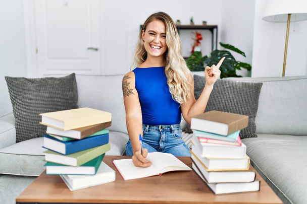 Young beautiful woman writing on notebook around university books smiling happy pointing with hand and finger to the side  - Photo, Image