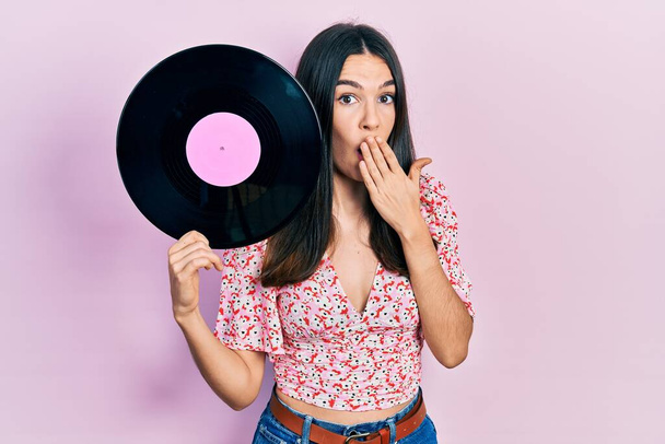 Young brunette woman holding vinyl disc covering mouth with hand, shocked and afraid for mistake. surprised expression  - Foto, afbeelding