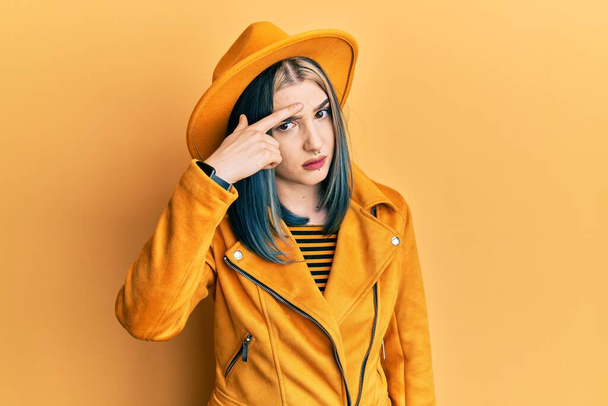 Young modern girl wearing yellow hat and leather jacket pointing unhappy to pimple on forehead, ugly infection of blackhead. acne and skin problem  - Foto, imagen