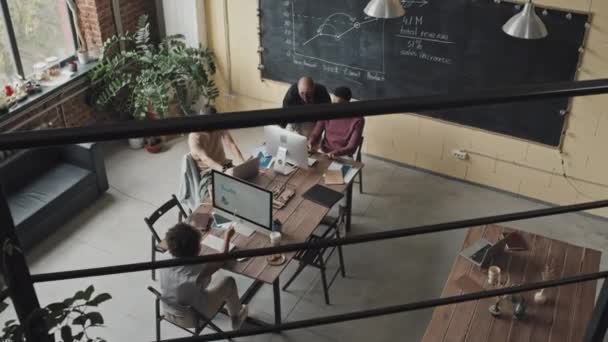 Hoge hoek van vier verschillende vrouwen en mannen met gezichtsmaskers, met behulp van computers, die overdag aan tafel in loft-stijl coworking ruimte zitten - Video