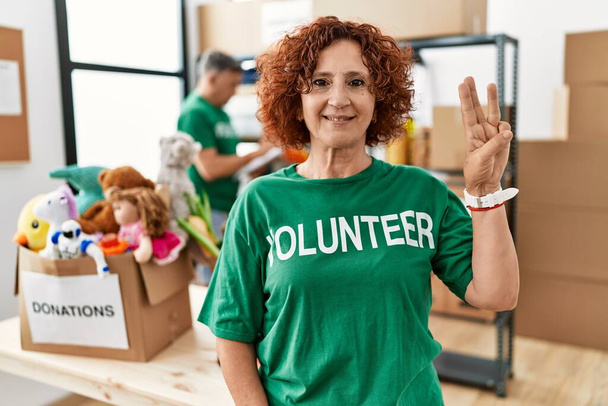 Egy középkorú nő, aki önkéntes pólót visel az adományoknál, mutogat és mutogat a hármas számú ujjaival, miközben magabiztosan és boldogan mosolyog..  - Fotó, kép