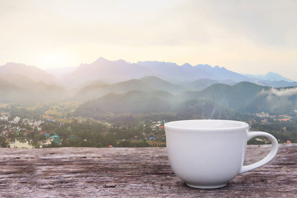Una taza blanca de tazas de café expreso caliente colocadas en un suelo de madera con niebla matutina y moutains con fondo de luz solar, café mañana - Foto, Imagen