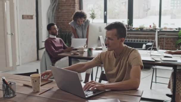 Derék-up fiatal férfi kaukázusi IT-szakember ül íróasztal forgalmas modern irodában nappal, laptoppal, gépelés - Felvétel, videó