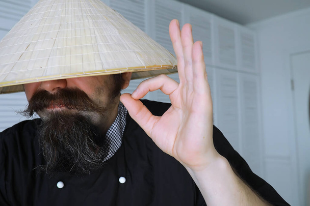 Un'esilarante parodia di un asiatico col cappello vietnamita con la barba. Ritratto. Asiatico caffè chef. - Foto, immagini
