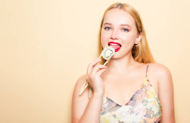 Girl in a dress on a beige background. Woman eating Philadelphia sushi. Blonde girl with red lipstick holding chopsticks for sushi and eating. - Photo, Image