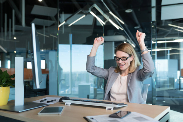 Beautiful and successful business woman with glasses, celebrating victory looks at the computer monitor screen and rejoices in the successfully completed project, keeps her hands up from happiness and satisfaction - Фото, изображение