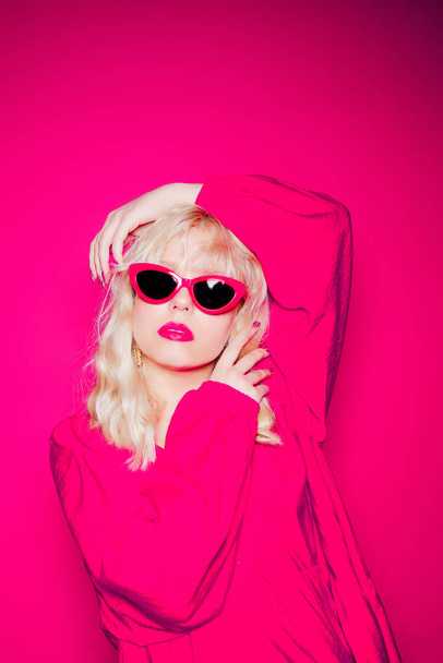 A szőke rózsaszín háttéren pózol elegáns rózsaszín szemüvegben és ruhában. Lány modell pózol, fényes fotó divat és stílus, arany ékszer 80-as és 90-es évek. - Fotó, kép